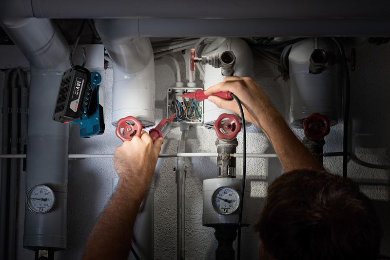 Person checking electrical wires near pipes, cordless work light attached above.