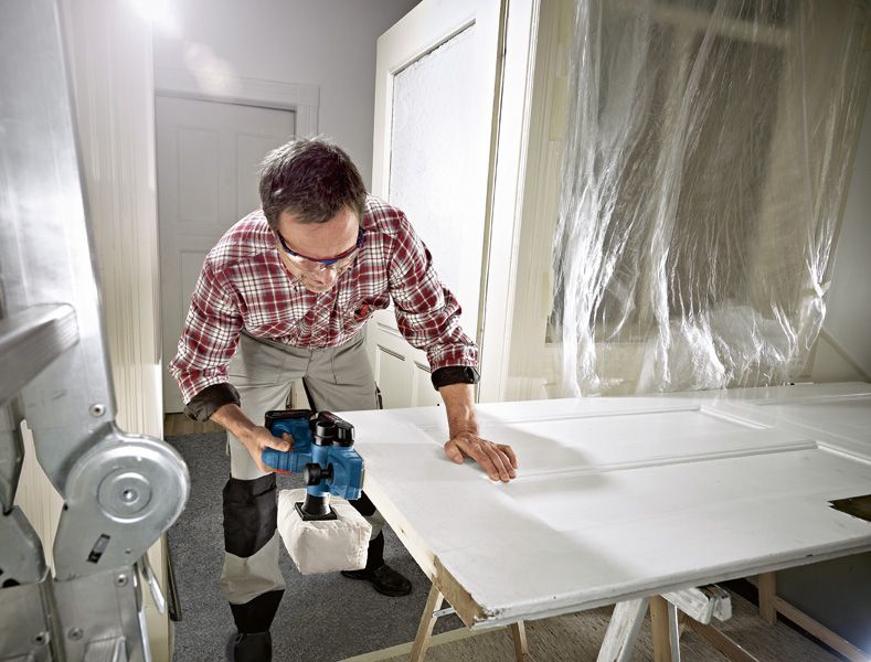 GHO 18V-LI A person wearing safety equipment uses a cordless planer to smooth a white door.