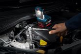 A person works on a car engine using a wrench with a cordless work light nearby.