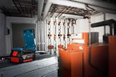 Cordless work light illuminates an industrial boiler room with pipes and orange machinery.