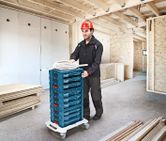 Person wearing safety equipment transports stacked tool cases on a wheeled cart in a building under construction.