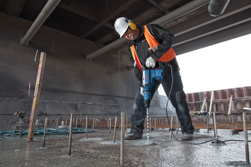 A person wearing safety equipment drills into concrete with a rotary hammer at a construction site.