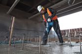 A person wearing safety equipment drills into concrete with a rotary hammer at a construction site.
