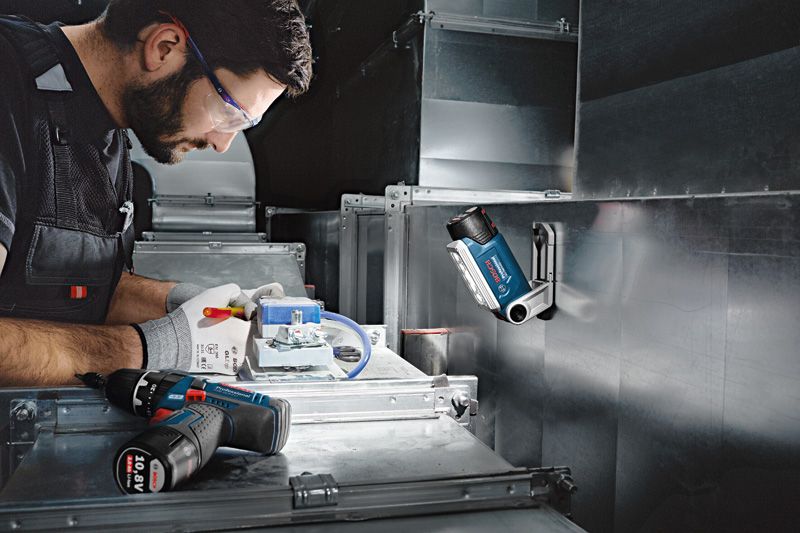 A person wearing safety equipment works with electrical wiring under a cordless work light.