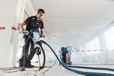 A person wearing safety equipment uses a demolition hammer with dust extraction on a concrete floor.