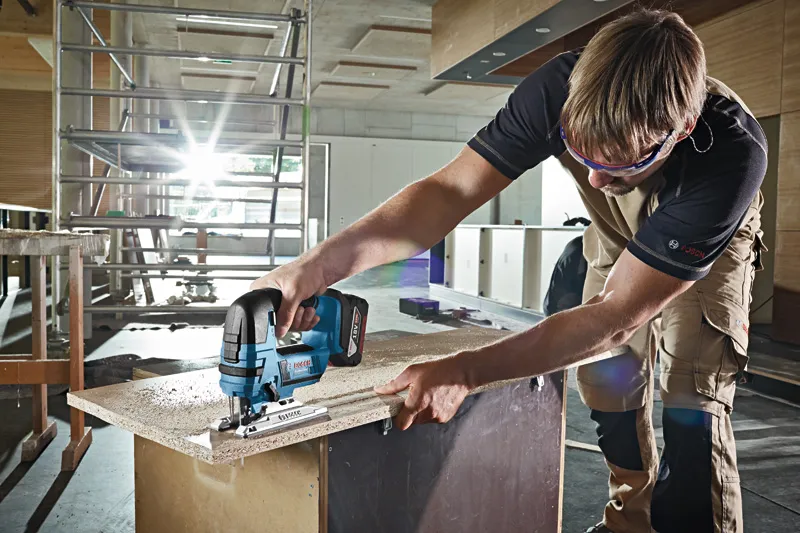 A person wearing safety equipment uses a cordless jigsaw to cut a wooden board.