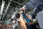 A person wearing safety equipment grinds a metal pipe with a cordless angle grinder.