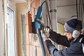 A person wearing safety equipment uses a wall chaser with a dust extraction hose on brick.