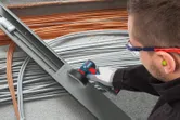 A person wearing safety equipment cuts a metal cable tray with a cordless angle grinder.