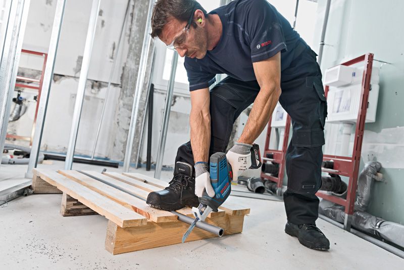 A person wearing safety equipment uses a reciprocating saw to cut a metal pipe.