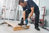 A person wearing safety equipment uses a reciprocating saw to cut a metal pipe.