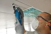 A person wearing safety equipment cuts a ceiling panel with a multi-cutter tool.