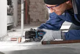 A person wearing safety equipment uses a multi-cutter to trim copper pipes on a dusty floor.