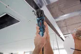 A person wearing safety equipment cuts a ceiling panel with a multi-cutter near metal framing.