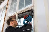 A person wearing safety equipment uses a reciprocating saw to cut a window frame.