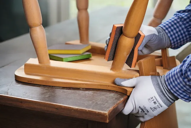 Person wearing safety equipment sands a wooden chair leg with a sanding sponge.