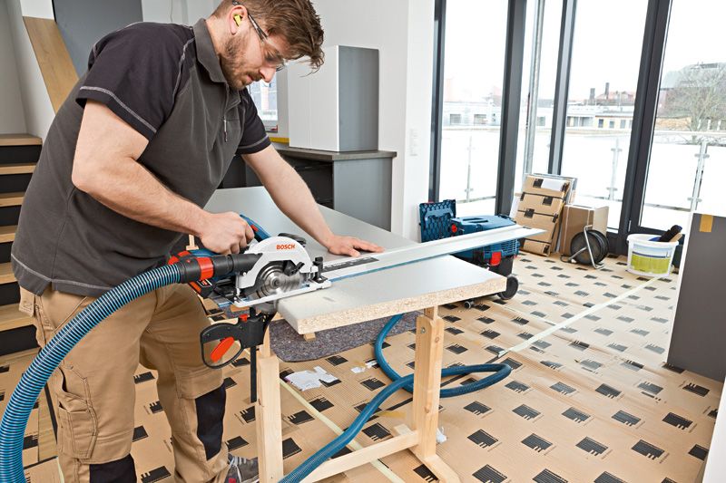 Person wearing safety equipment cuts wood panel with a cordless circular saw on a workbench.