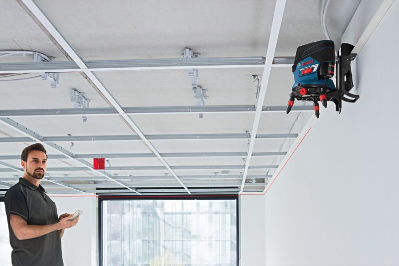 A person operates a laser leveling tool mounted on a wall to align ceiling framework.