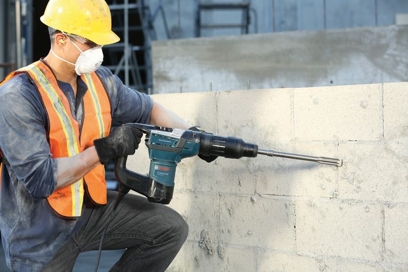 A person wearing safety equipment uses a rotary hammer to drill into a concrete wall.