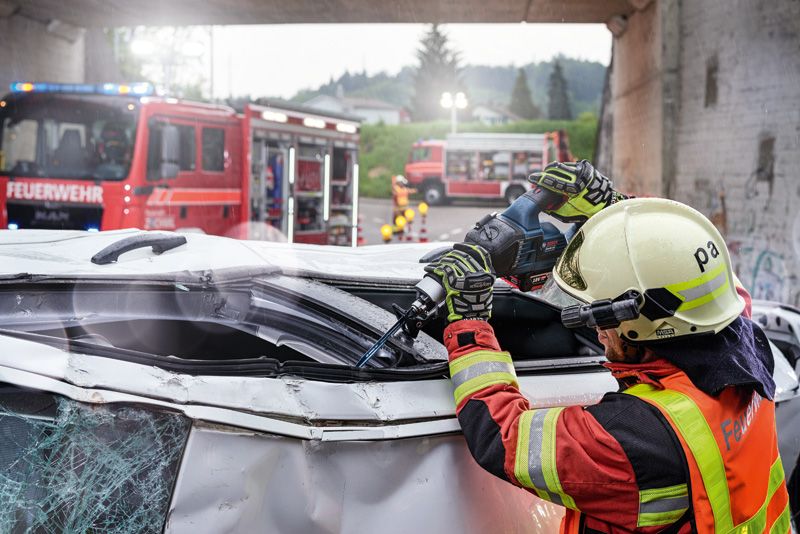 A person wearing safety equipment uses a power saw to cut a damaged vehicle roof.
