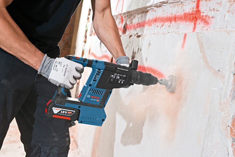A person wearing safety equipment drills into a concrete wall with a cordless rotary hammer.