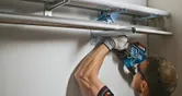 Person wearing safety equipment uses a power drill to mount metal pipes on a wall.
