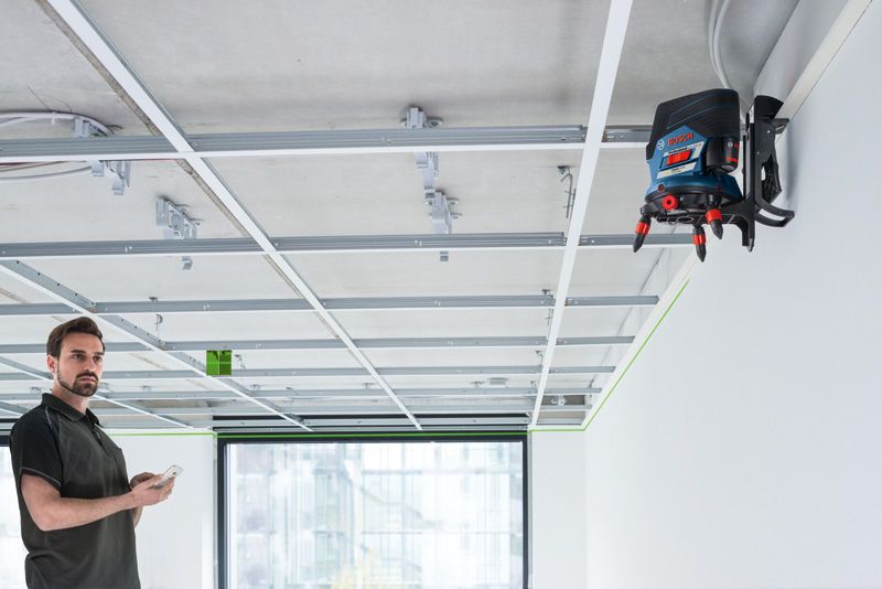 GCL 2-50 CG Person using a phone observes a laser leveling tool projecting a green line on a suspended ceiling frame.