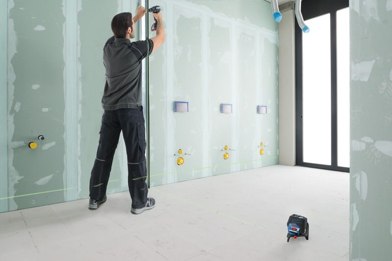 GCL 2-50 CG Person adjusting pipe on unfinished wall, green laser leveling tool projecting horizontal line.