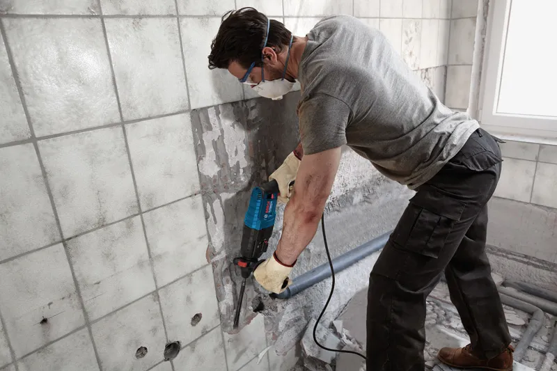 GBH 2-24 DRE A person wearing safety equipment uses a power tool to remove tiles from a wall.