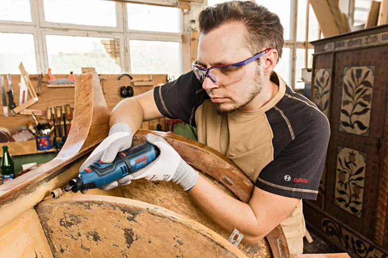 A person wearing safety equipment sands wood with a cordless rotary tool in a workshop.
