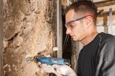 A person wearing safety equipment grinds a wall using a cordless rotary tool.