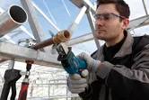 A person wearing safety equipment uses a cordless reciprocating saw to cut a metal pipe.