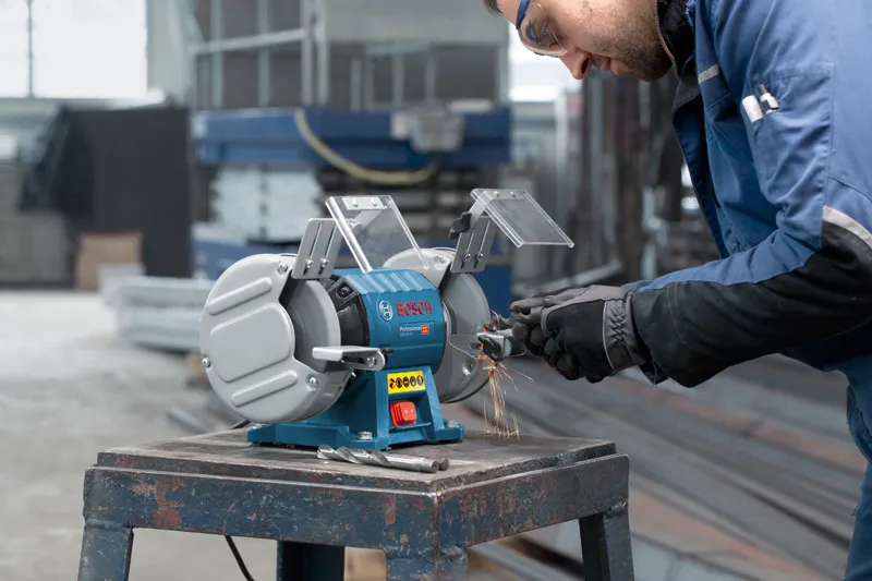 A person wearing safety equipment grinds metal on a double-wheeled bench grinder.
