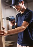 Person wearing safety equipment drills a hole into a wooden board.