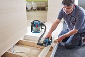 A person wearing safety equipment uses a cordless planer on wooden boards indoors.