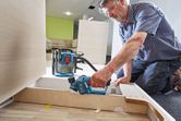 A person wearing safety equipment uses a cordless planer to smooth wooden panels.