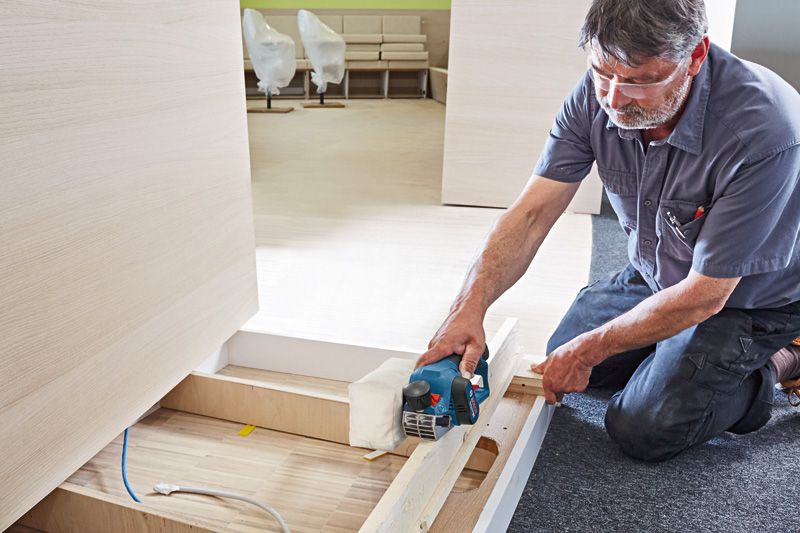 GHO 12V-20 A person uses a cordless planer to smooth a wooden board on a jobsite floor.