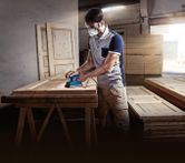 A person wearing safety equipment sands a wooden door with an orbital sander in a workshop.