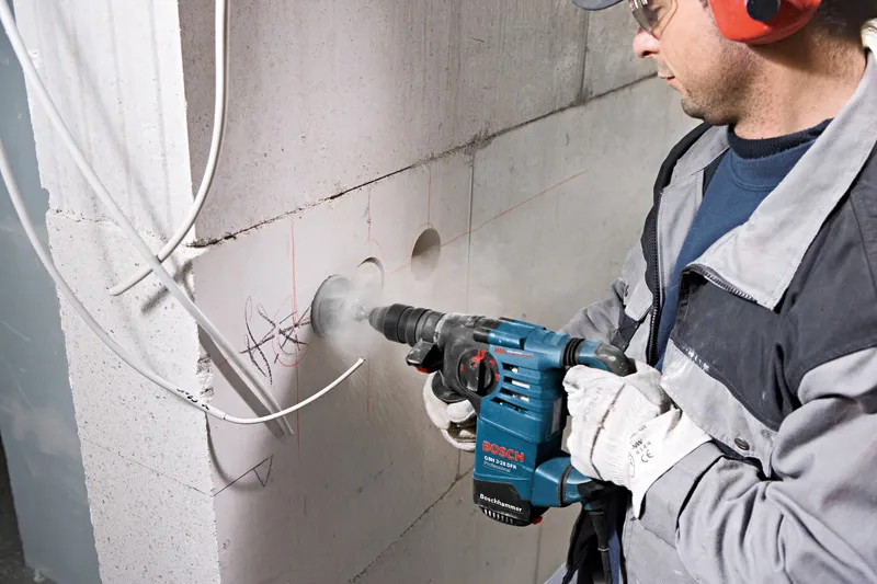 A person wearing safety equipment drills into a concrete wall with a rotary hammer.