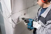 A person wearing safety equipment drills into a concrete wall with a rotary hammer.