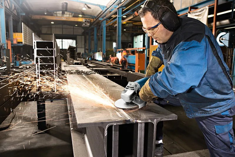 A person wearing safety equipment grinds a large metal beam with an angle grinder.