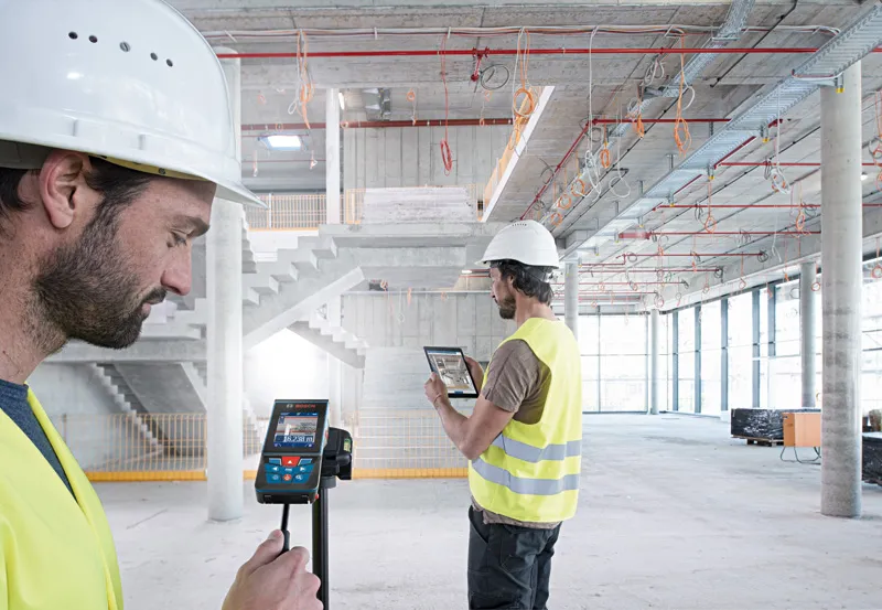 GLM 150 C Two people wearing safety equipment use a laser measure and tablet inside a large construction site.