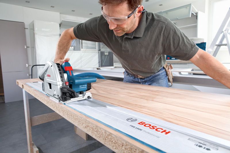 A person wearing safety equipment uses a plunge saw to cut wood along a guide rail.