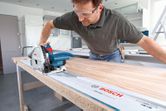 A person wearing safety equipment uses a plunge saw to cut wood along a guide rail.