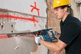 A person wearing safety equipment chisels a groove into a marked concrete wall.