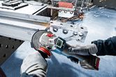 A person wearing safety equipment grinds metal, producing sparks with a cordless angle grinder.