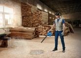 A person wearing safety equipment uses a blue blower to clear sawdust in a woodshop.