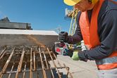 A person wearing safety equipment cuts rusty rebar with an angle grinder at a construction site.