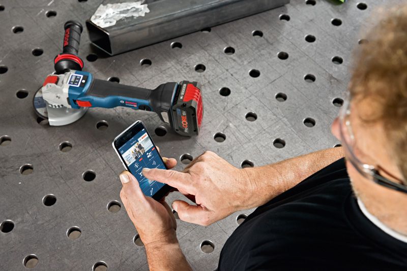 A person wearing safety equipment uses a smartphone to control a cordless angle grinder.