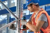 A person wearing safety equipment secures a metal frame with a cordless power drill.